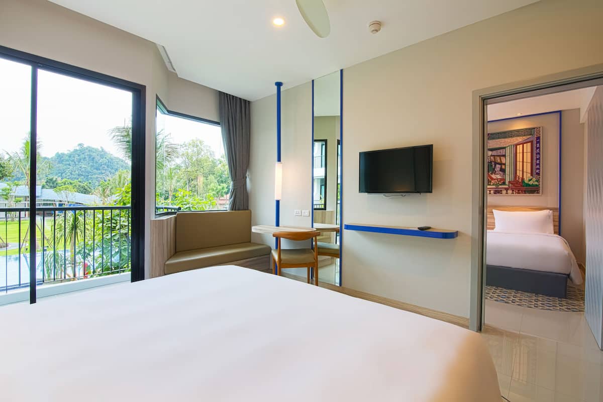 A hotel room with a large bed, a TV mounted on the wall, a balcony with a view of greenery, and a small table with a chair.