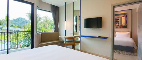 Quarto de hotel com uma cama grande, televisão presa à parede, varanda com vista para áreas verdes e uma pequena mesa com uma cadeira.