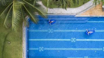 An aerial view of a swimming pool with two inflatable rings, surrounded by palm trees and a grassy area.