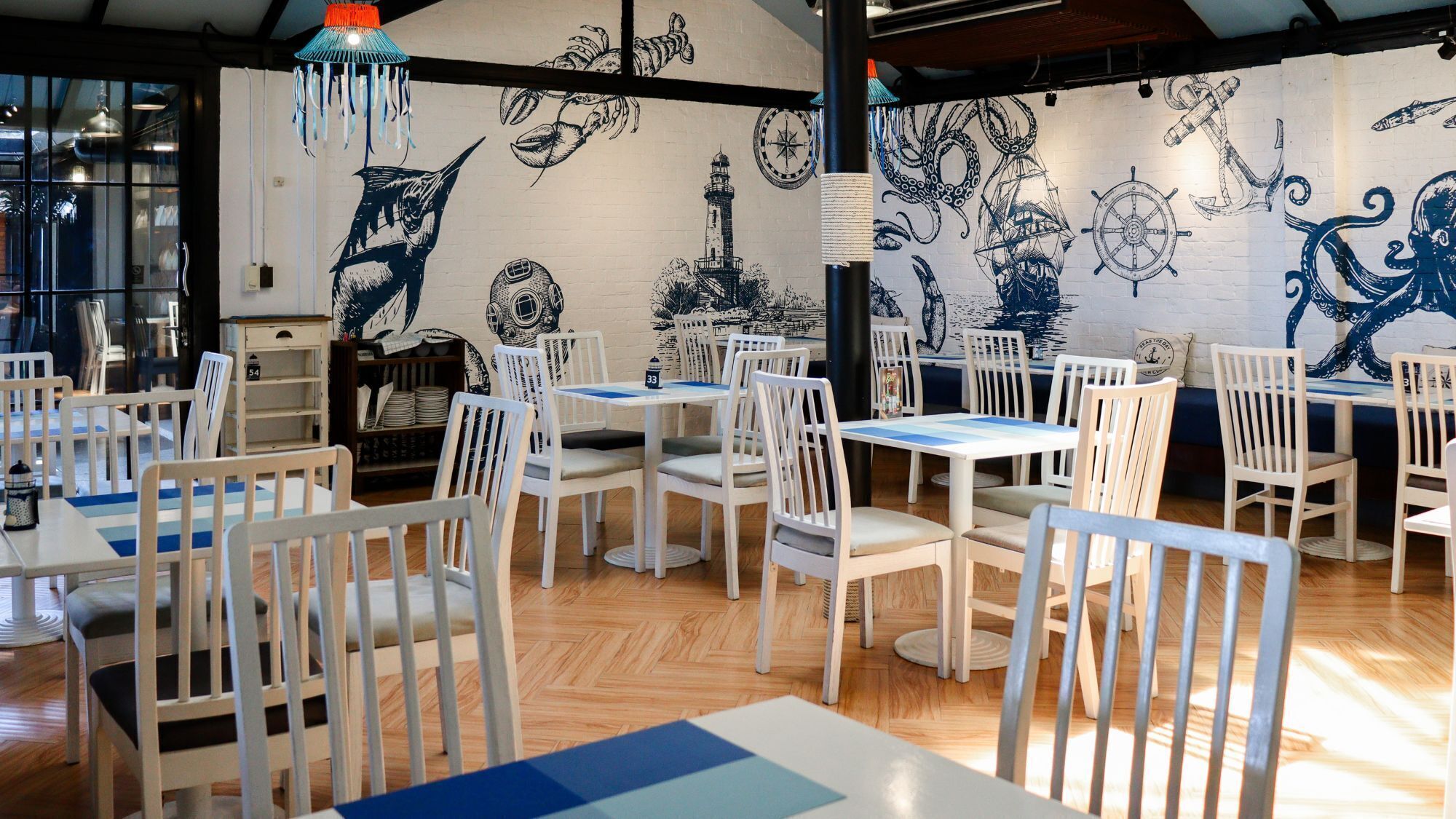 A restaurant with white chairs and tables, nautical-themed wall art, and a wooden floor.