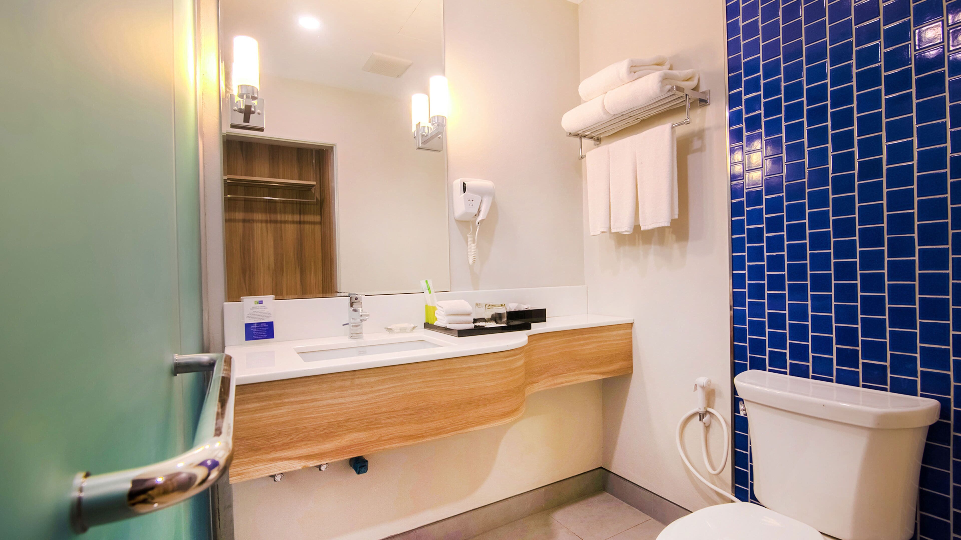 Un bagno con un mobile vanitario in legno, un lavandino bianco, uno specchio, un portasciugamani e una parete rivestita di piastrelle blu.