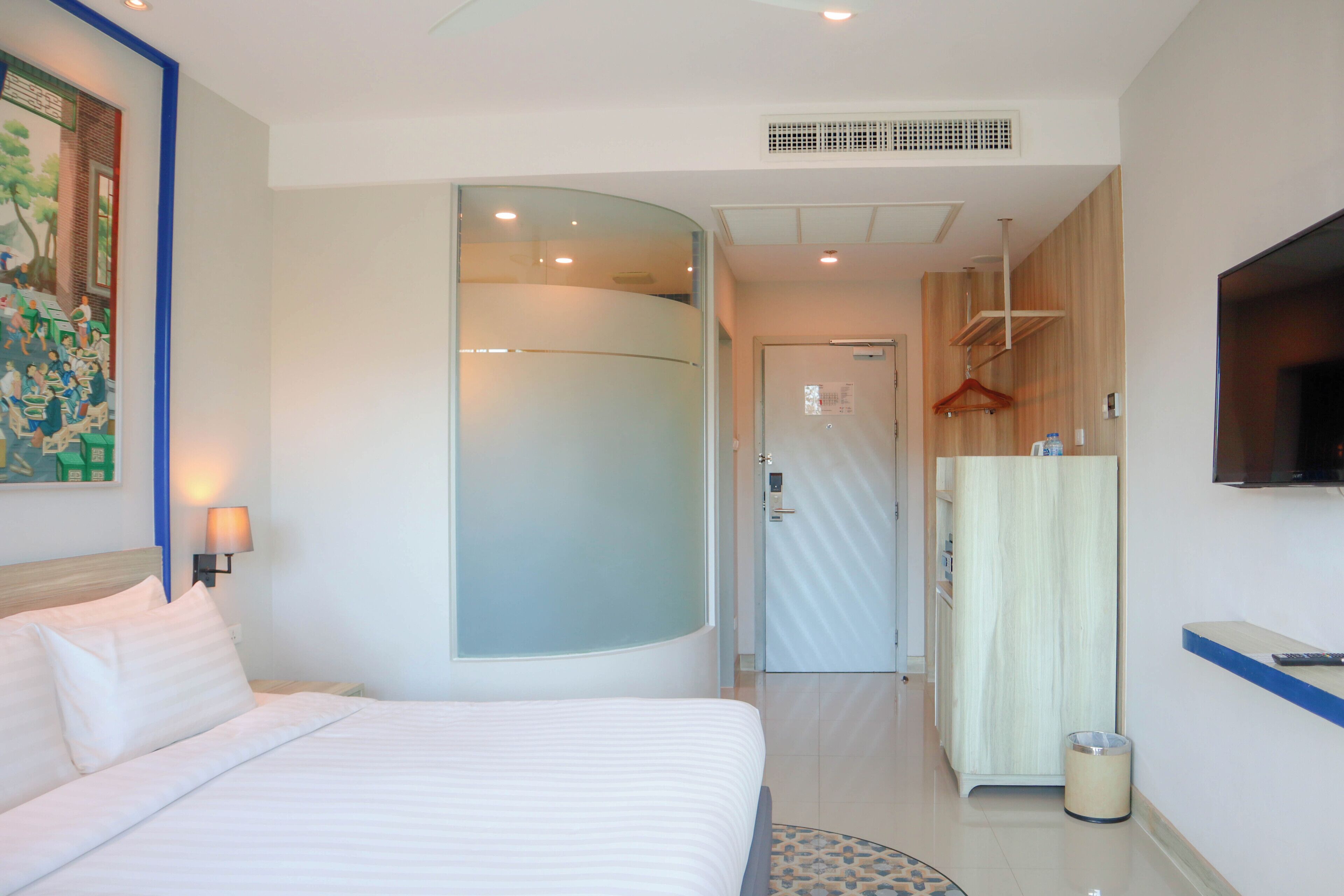 A hotel room with a bed, a TV mounted on the wall, a mirror, a coat rack, and a waste bin.