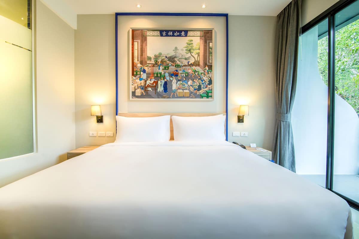 A hotel room with a large bed, two bedside tables with lamps, a wall-mounted painting, and a large window with curtains.