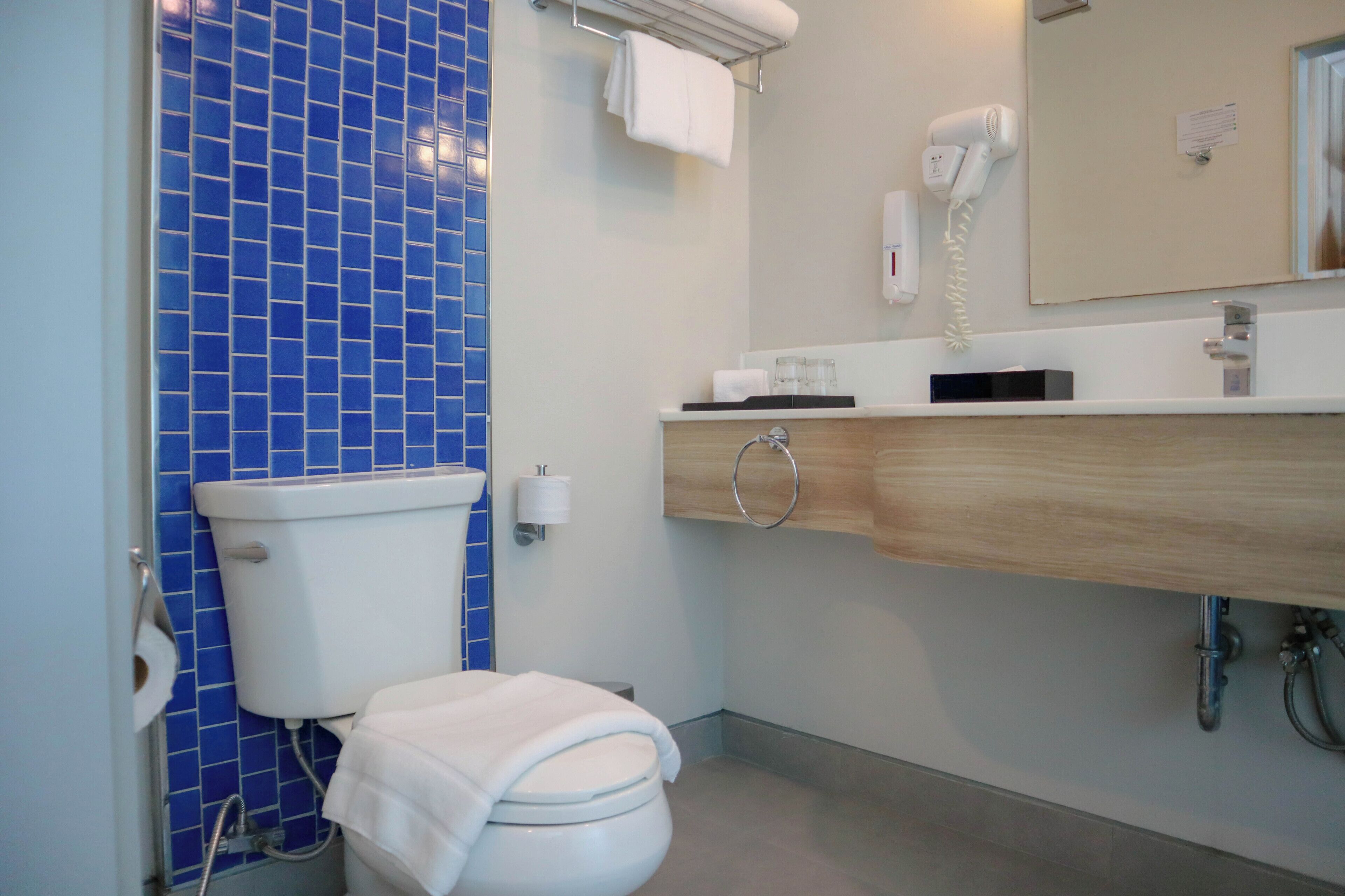 A bathroom with a blue-tiled wall, a white toilet, a sink with a wooden countertop, a mirror, and a hairdryer on the wall.