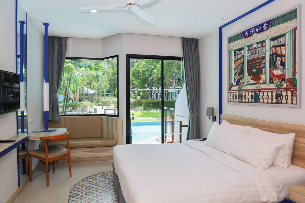 A hotel room with a bed, a desk, a chair, a television, and a large window with a view of a pool and greenery.