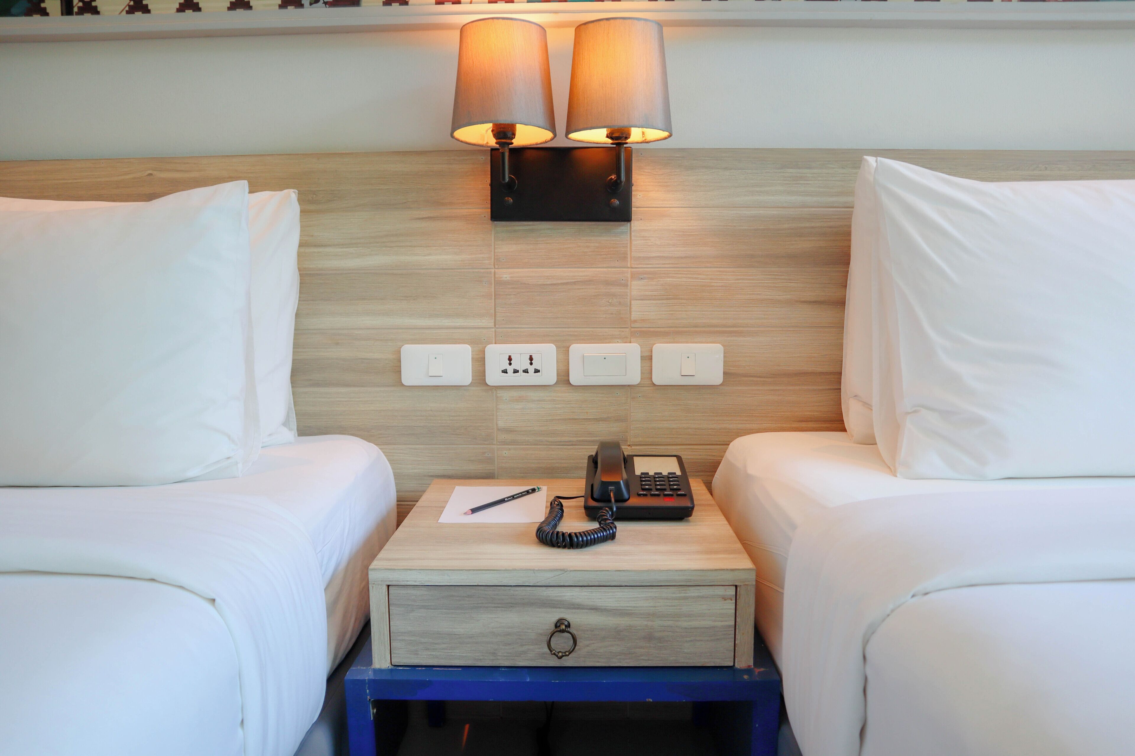 A hotel room with two beds, a bedside table with a lamp, a phone, and a drawer.