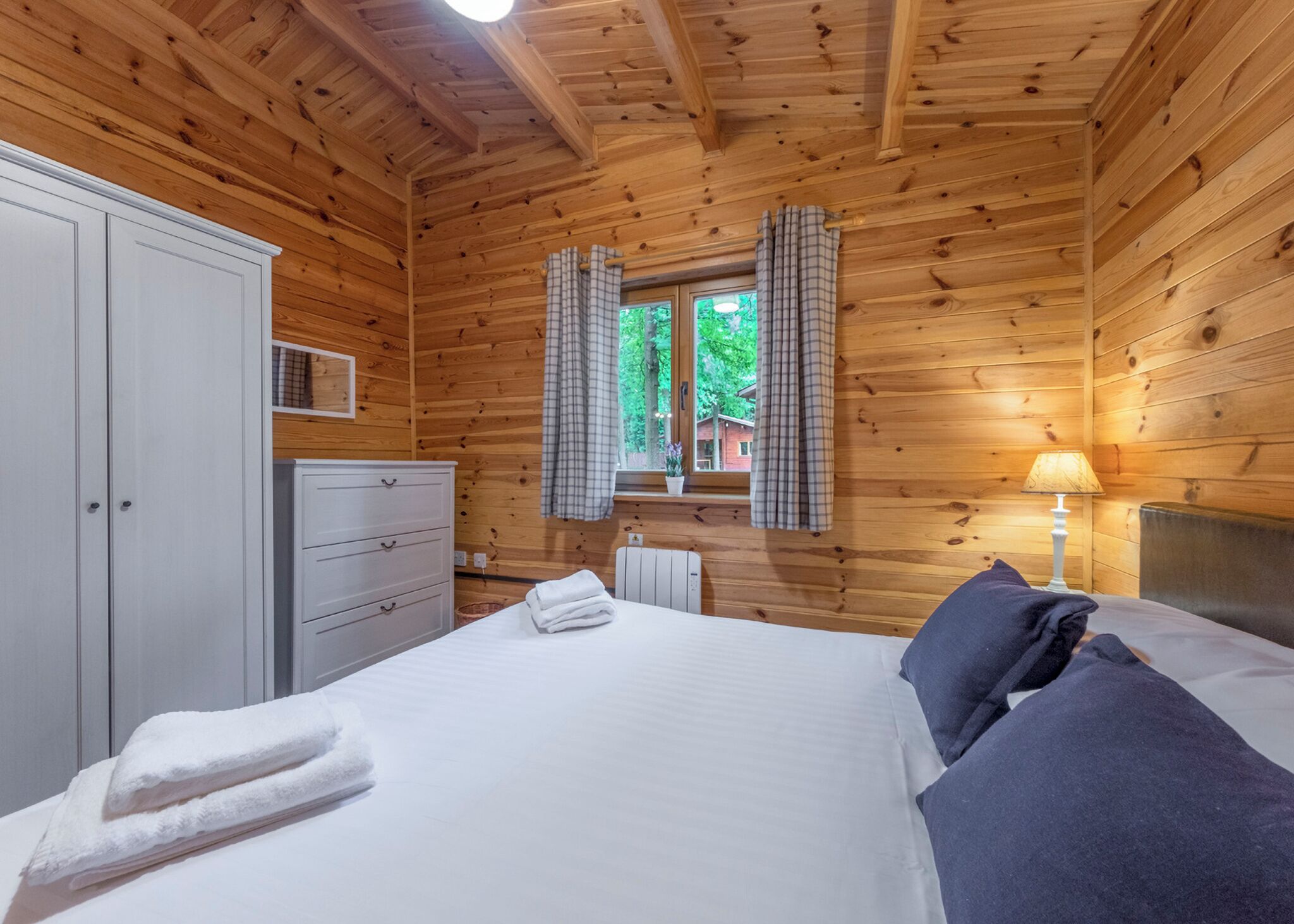 A wooden cabin bedroom with a bed, a wardrobe, a bedside table with a lamp, and a window with curtains.