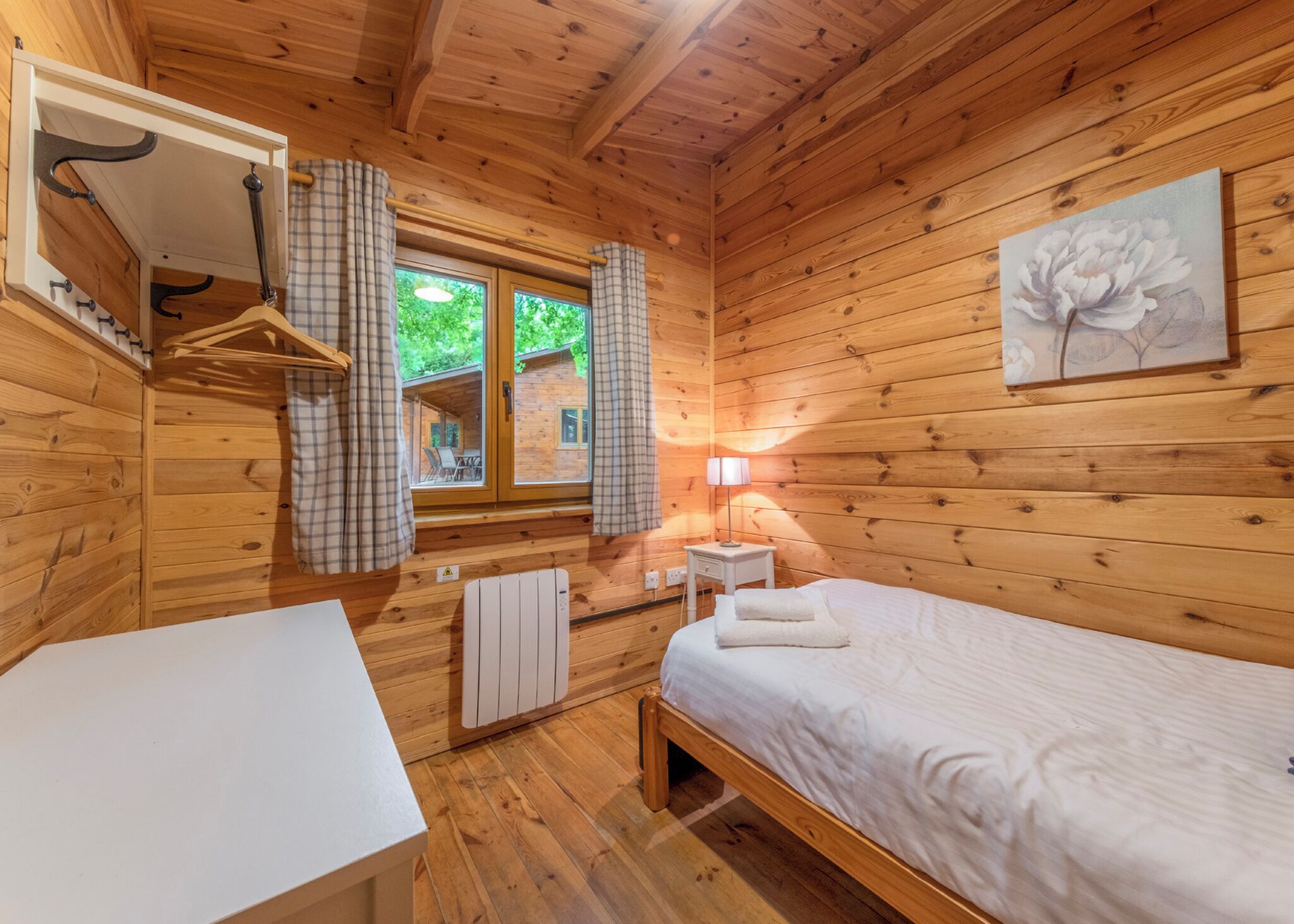 A wooden cabin room with a bed, a table, a window with curtains, a radiator, and a wall-mounted lamp.