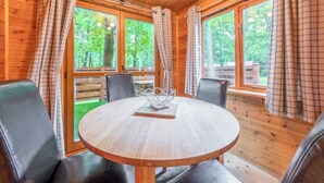 A wooden cabin interior with a round table, four chairs, plaid curtains, and large windows.