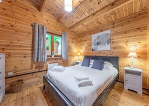 Une chambre à coucher aux murs lambrissés, avec un lit, deux tables de chevet dotées de lampes, une fenêtre avec des rideaux et un tableau représentant une orchidée accroché au mur.