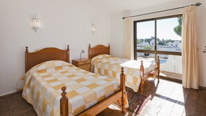 A room with two wooden beds, a nightstand, a lamp, and a view of a building through a large window. - Casa ADM 42 - Villa with Pool, Walk to Carvoeiro (Lagoa)