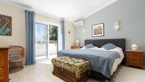 Un dormitorio con una cama, un aparador, una silla de mimbre y vistas a través de un ventanal.