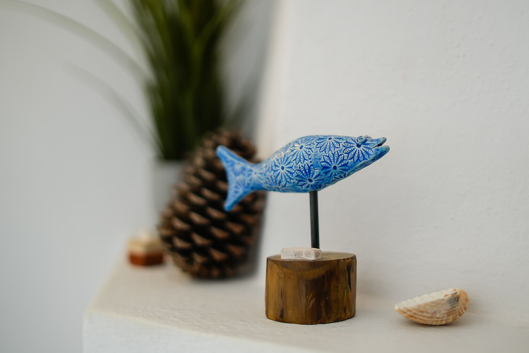 Een blauwe decoratieve vis op een houten standaard, met een dennenappel en schelpen op de achtergrond.