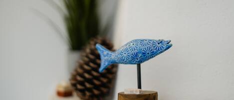 Een blauwe decoratieve vis op een houten standaard, met een dennenappel en schelpen op de achtergrond.