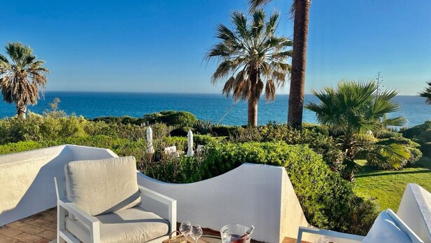 A terrace with white furniture, a table with drinks, and a view of the sea and palm trees.