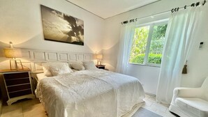 A bedroom with a large bed, a nightstand, a chair, a window with curtains, and a painting on the wall.