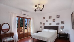 Een slaapkamer met een groot bed, een houten dressoir, een spiegel, een nachtkastje met een lamp, en uitzicht door een glazen deur op een buitenruimte.