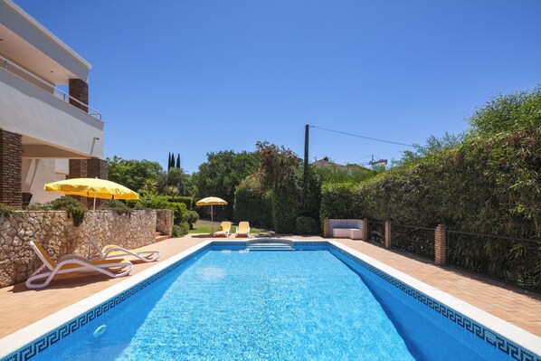 A swimming pool with two sun loungers and umbrellas, surrounded by a stone wall and greenery.