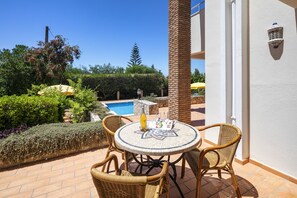 Une terrasse avec des chaises et une table en osier, une piscine en arrière-plan et un ciel bleu dégagé.