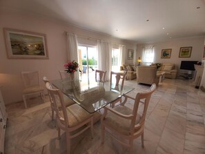 A spacious living area with a glass dining table surrounded by chairs, a sofa, and a television.