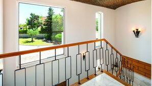 Una escalera con barandilla de madera y balaustres de metal negro, que conduce a un rellano con un gran ventanal que ofrece vistas a la vegetación y un edificio.