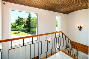 Eine Treppe mit Holzhandlauf und schwarzen Metallspindeln, die zu einer Aussichtsplattform mit einem großen Fenster führt, das Blick auf Grünflächen und ein Gebäude gewährt.