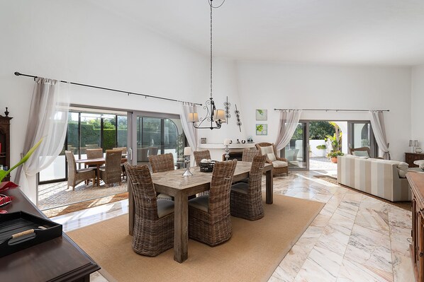 A spacious dining area with a wooden table and wicker chairs, a chandelier, and a view through large windows. - Casa Blanca, 3 bedroom villa with air conditioning and private pool (Carvoeiro)