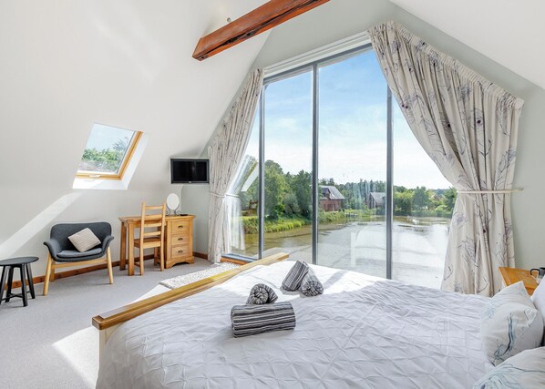 A bedroom with a large bed, a desk, a chair, a TV, and a view of a river. - Stunning two storey contemporary lodge on the edge of the lake. (Anglesey)