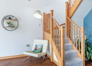 A staircase with wooden railings, a cushioned chair with a colorful pillow, a pendant light, and a wall-mounted shelf with plants. - Stunning two storey contemporary lodge on the edge of the lake. (Anglesey)
