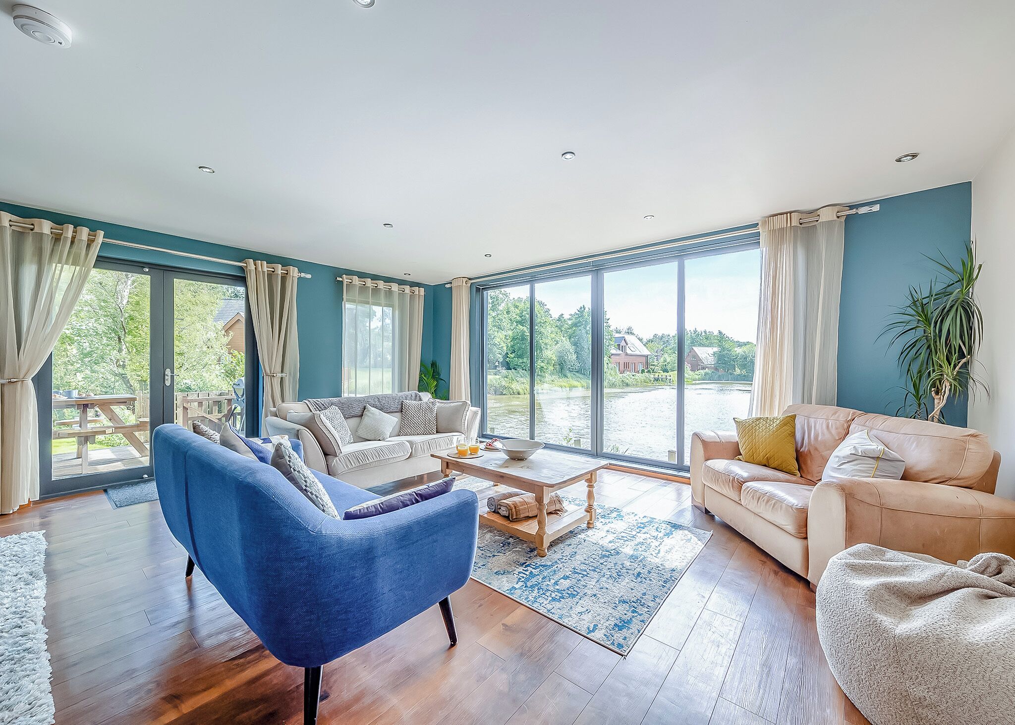 A spacious living room with a blue armchair, two sofas, a coffee table, and large windows offering a view of a lake and greenery.
