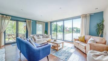 A spacious living room with a blue armchair, two sofas, a coffee table, and large windows offering a view of a lake and greenery.