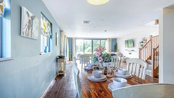 A dining area with a wooden table set for four, surrounded by white chairs, with artwork on the walls and a staircase leading to another level.