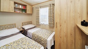 A room with two beds, a wardrobe, a shelf with items, and a window with curtains. - A modern holiday home with one master double and two twin bedrooms. (Cresswell beach)