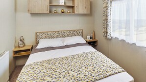 A bedroom with a bed, a nightstand, a dresser, a clock, and a window with curtains. - A modern holiday home with one master double and two twin bedrooms. (Cresswell beach)