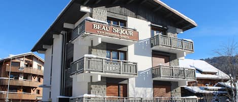 A multi-story hotel with a sign reading "BEAU SEJOUR" and a website address. It features balconies with wooden railings and a stone facade.