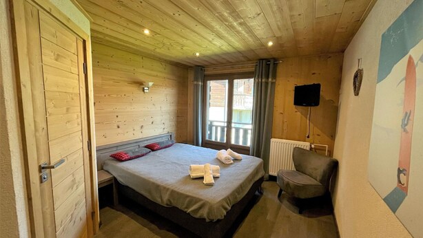 Un dormitorio con paneles de madera, una cama, una silla, un televisor y una ventana con cortinas.