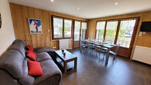 Una sala de estar acogedora con un sofá, una mesa de comedor y sillas. Hay ventanas amplias con cortinas, un radiador y un televisor montado en la pared.