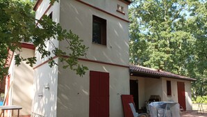 A building with a red door and windows, a covered patio area with a table and chairs, and a brick pathway leading to the entrance. - Country house in the heart of the Landes de Gascogne (Caubeyres)