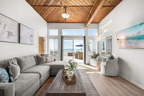 Ein großzügiges Wohnzimmer mit Holzboden, großen Fenstern und Blick auf den Strand.
