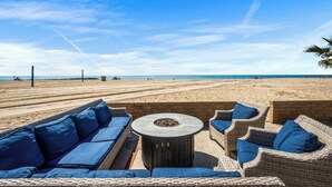 A beachside lounge with wicker furniture and a central fire pit, overlooking the beach and ocean.