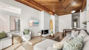 A spacious living room with a wooden ceiling, a fireplace, and a surfboard decoration.