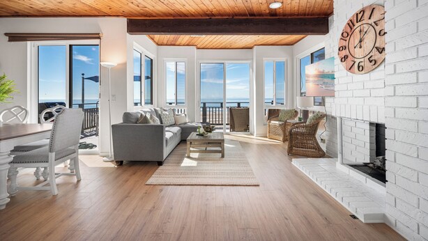 A spacious living room with a wooden ceiling, a fireplace, and large windows offering a view of the sea. - 3 Bedroom Oceanfront Home on 41st St (Newport Beach)