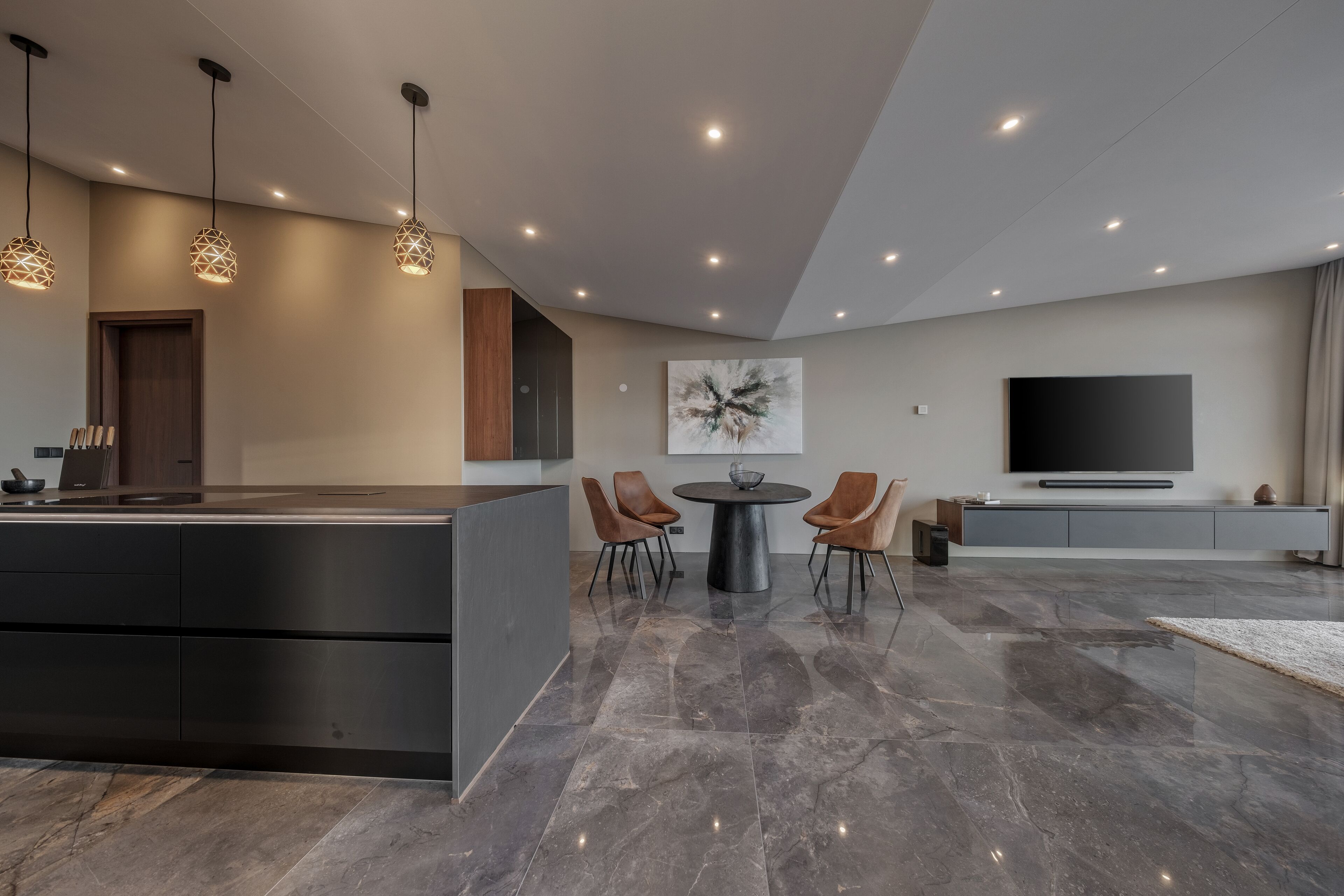 A modern kitchen with a central island, pendant lighting, a dining area with chairs, a large flat-screen TV, and a marble floor.