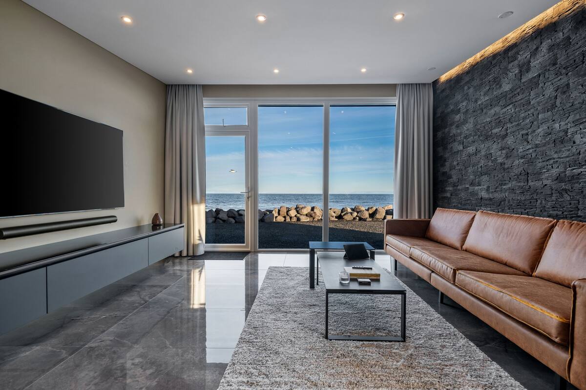 Uma sala de estar moderna com uma televisão de tela plana grande, um sofá de couro, uma mesa de centro e vista para o mar através de janelas do chão ao teto.