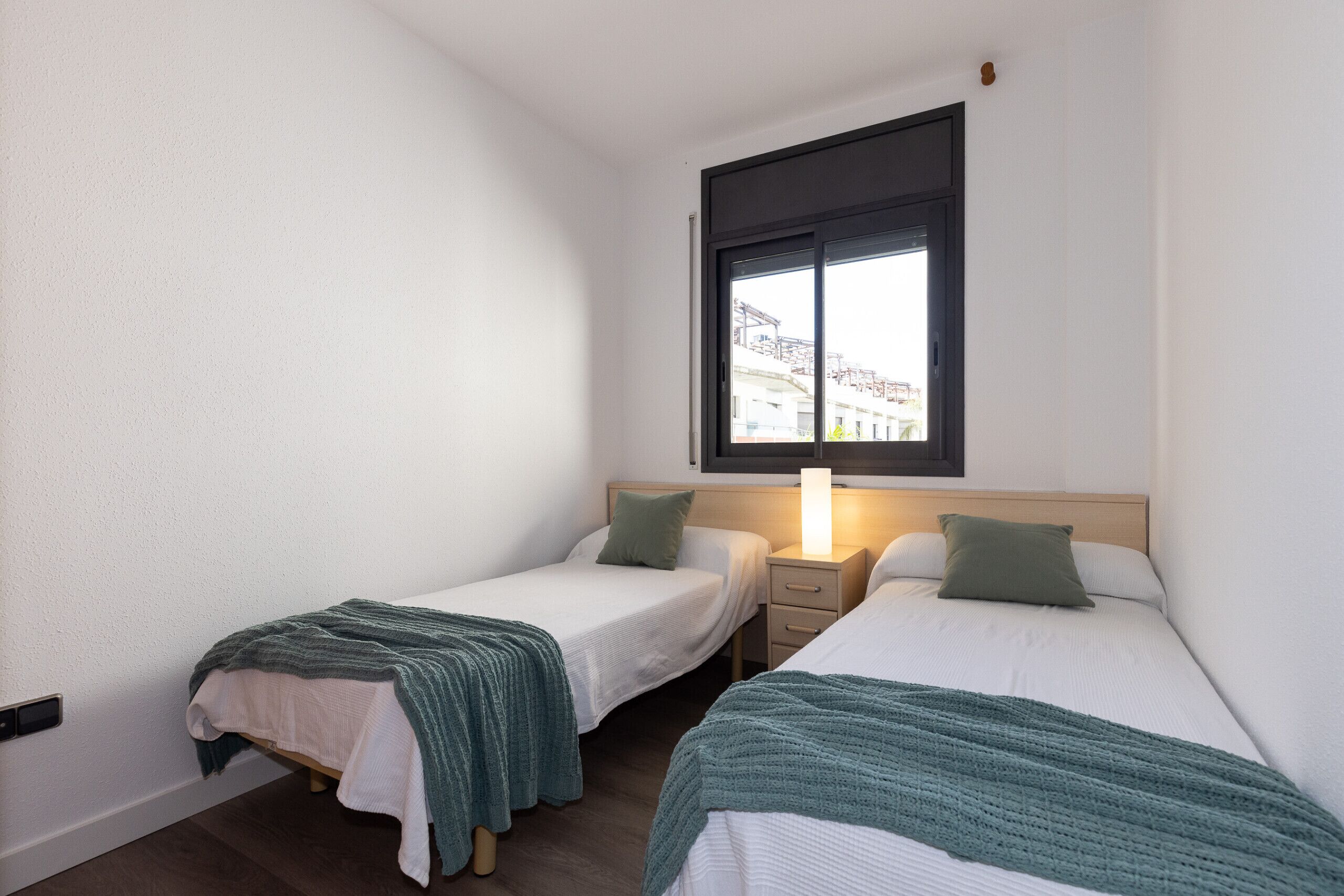 Una habitación pequeña y limpia con dos camas individuales, una mesita de noche, una lámpara y una ventana con vistas a edificios.