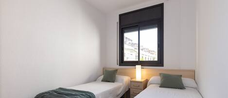 Una habitación pequeña y limpia con dos camas individuales, una mesita de noche, una lámpara y una ventana con vistas a edificios.