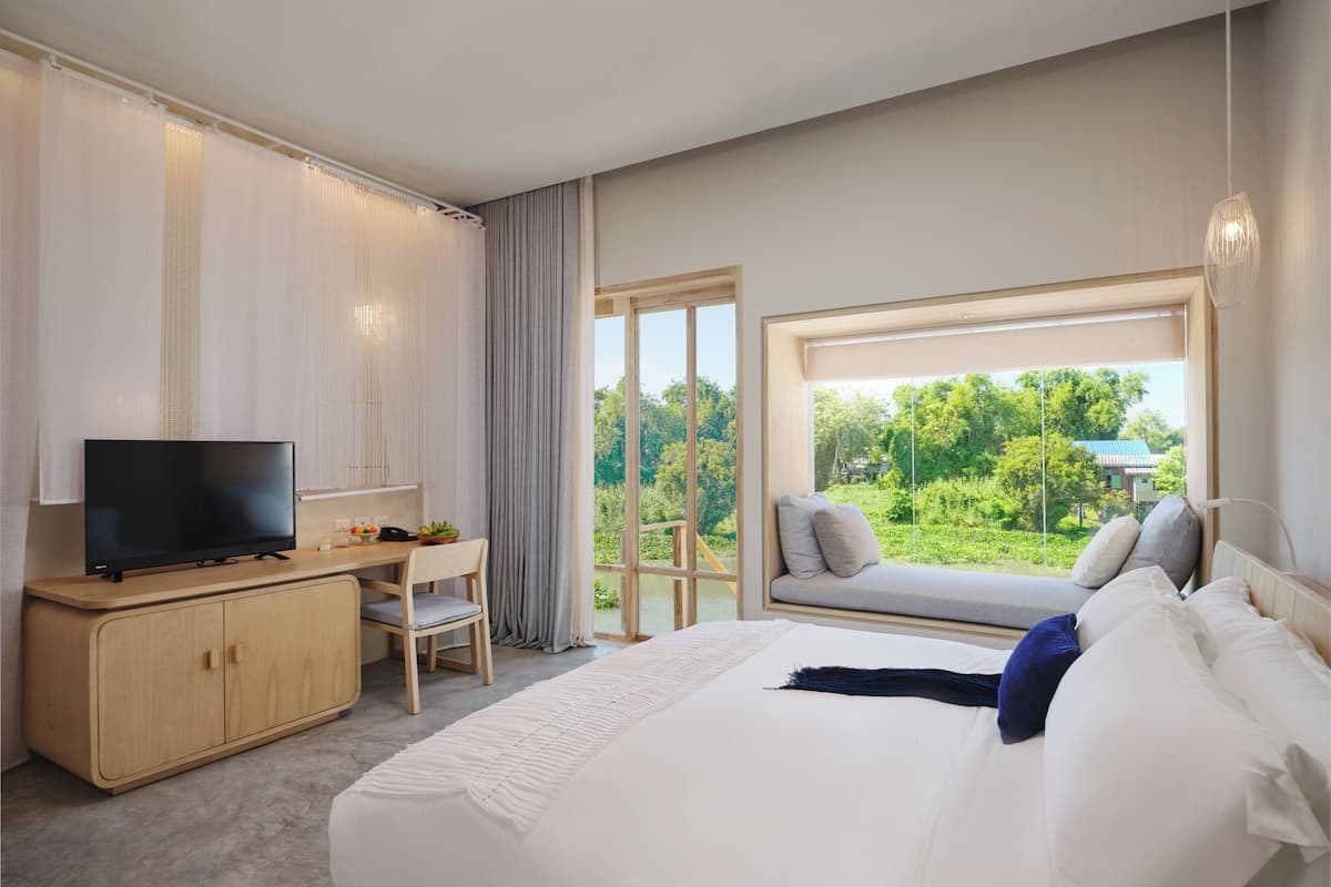 A modern hotel room with a large bed, a desk with a television, and a view of greenery outside.