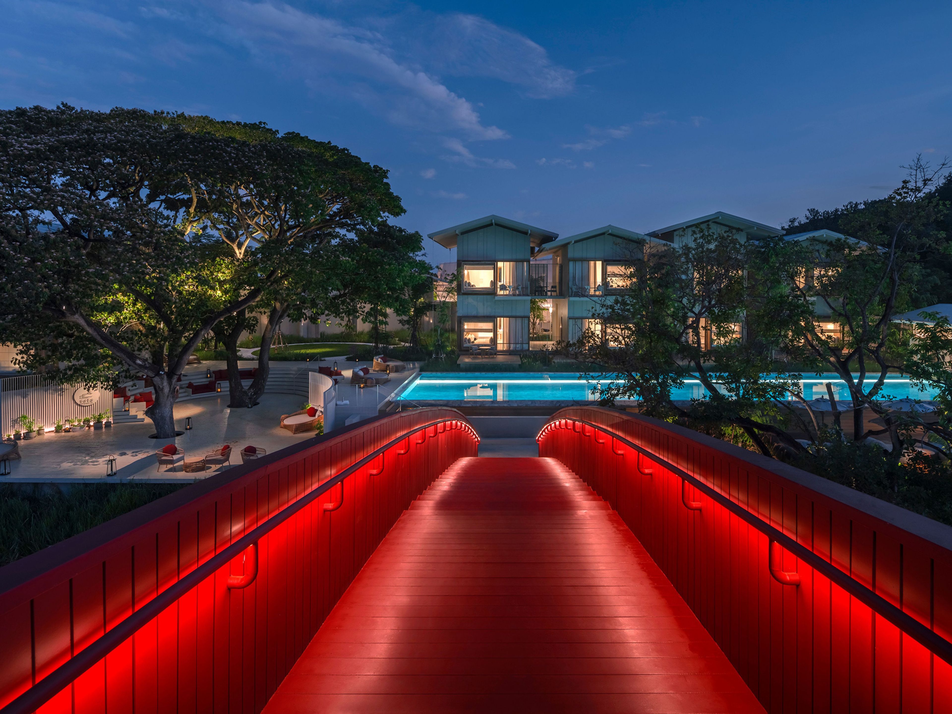 Ein modernes Hotel mit Schwimmbad, einer rot beleuchteten Brücke und üppigem Grün.