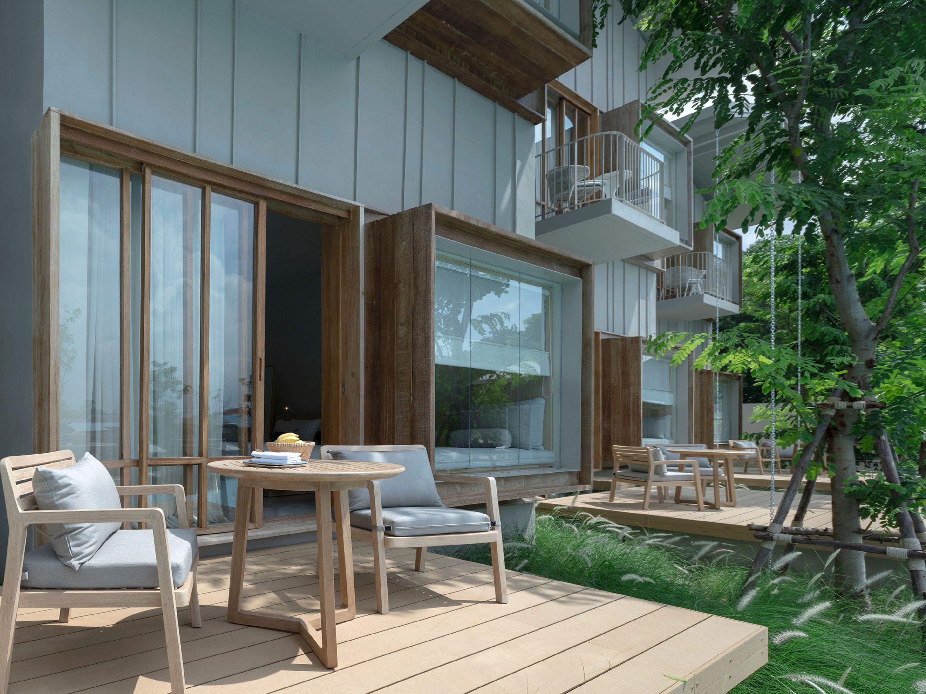 Un área de estar moderna al aire libre con mobiliario de madera, un acceso por puerta de vidrio y un balcón con barandilla blanca.