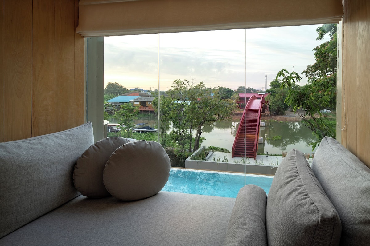 A room with a view of a swimming pool, a red building, and greenery.